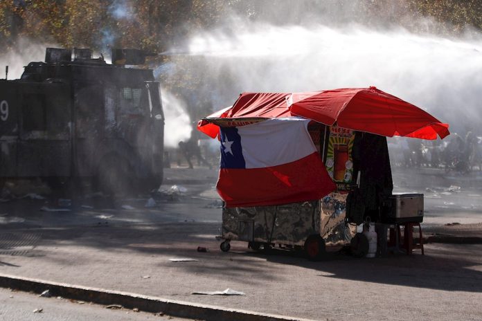 Un puesto callejero de venta de comida pasa frente a un vehículo lanza agua de las Fuerzas Especiales de Carabineros el viernes 7 de febrero, durante una nueva jornada de protestas en la plaza Italia, en Santiago. | Foto: Efe.